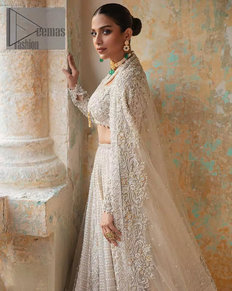 With a touch of Divine. A stunning ivory blouse is laboriously frosted with meticulously detailed hand embellishment with kora, dabka, tilla and pearls all over. The sweetheart neckline of the blouse and full sleeves together gives a so romantic look to any bride. In addition to this, it is systemized with the same colour frilled lehenga which is laden with the same colour embroidery that gives a magical stream of tilla, dabka and pearls. Finish this ivory article with the same colour dupatta that is heavily embellished with four-sided handsome borders and sequins sprayed all over to enhance the feeling of joy. 